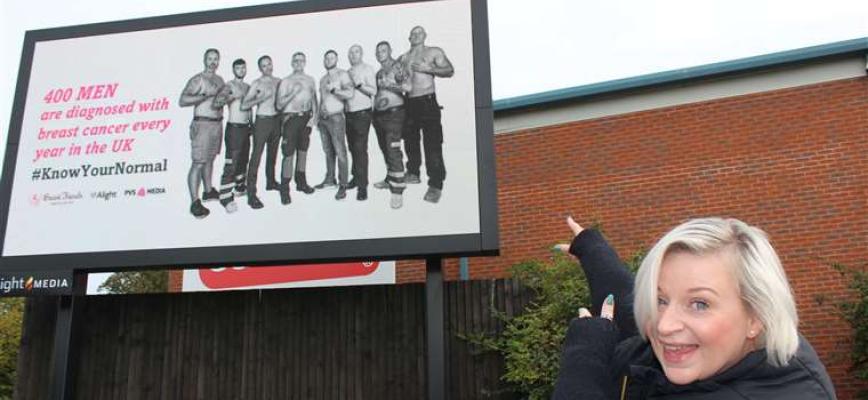 Rachael Bradley pictured smiling and pointing towards the billboard, featuring topless men with a health message. 